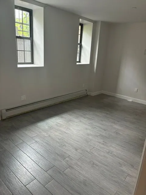 Yonkers Ave #1 bright room with two windows, gray flooring, and baseboard heating